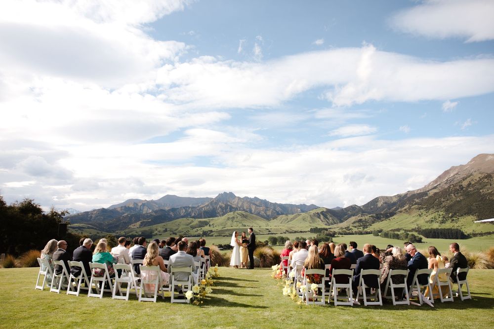 Outdoor wedding ceremony on lush green lawn with mountain backdrop and blue sky in scenic valley setting.