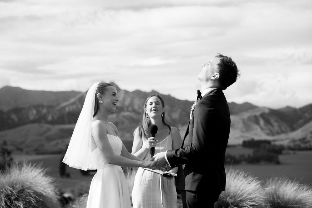 Black and white photo of bride and groom laughing with officiant during outdoor wedding ceremony with mountain backdrop.