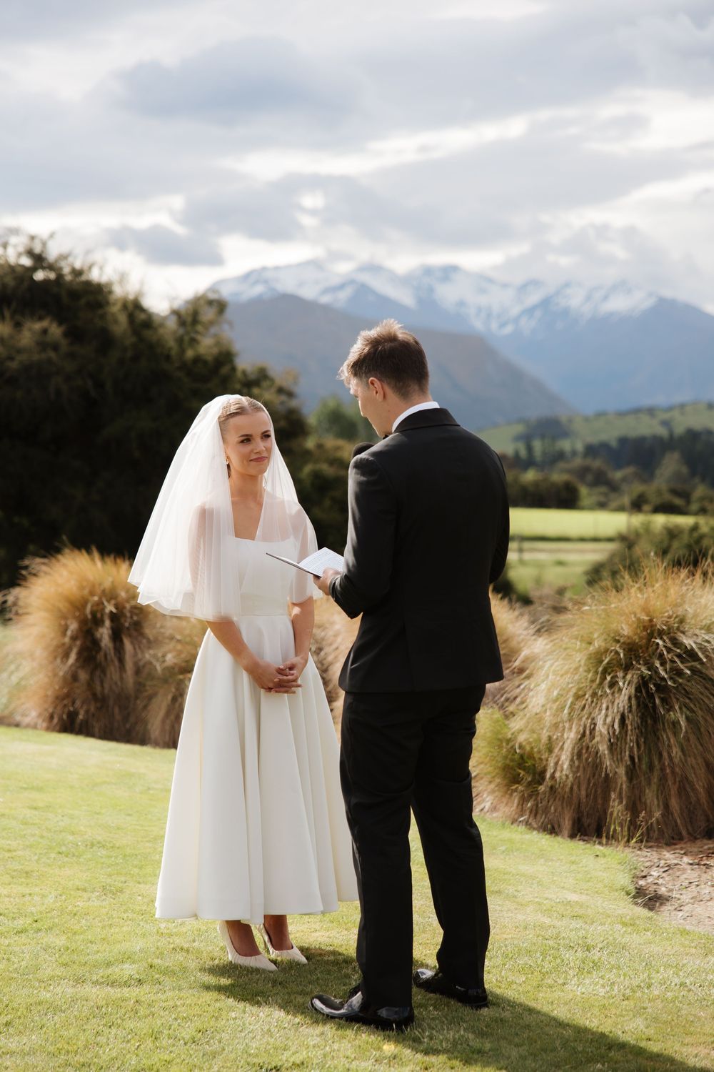 Bride and groom exchanging vows outdoors with snow-capped mountains and lush greenery behind them.