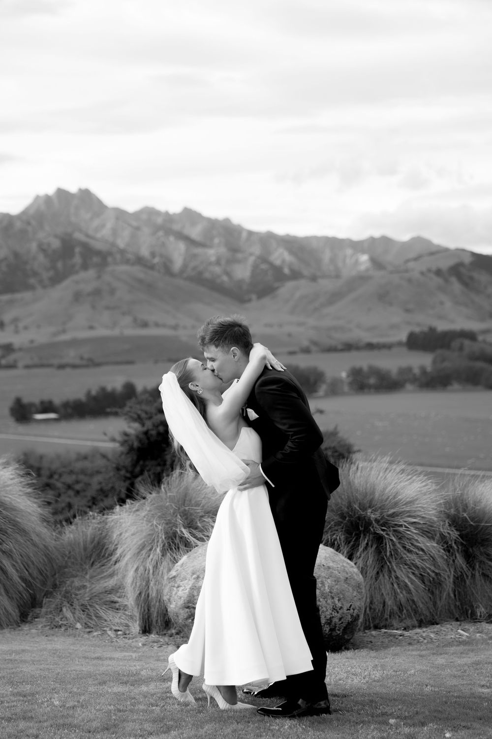 Bride and groom share romantic kiss with stunning mountain landscape backdrop in black and white.