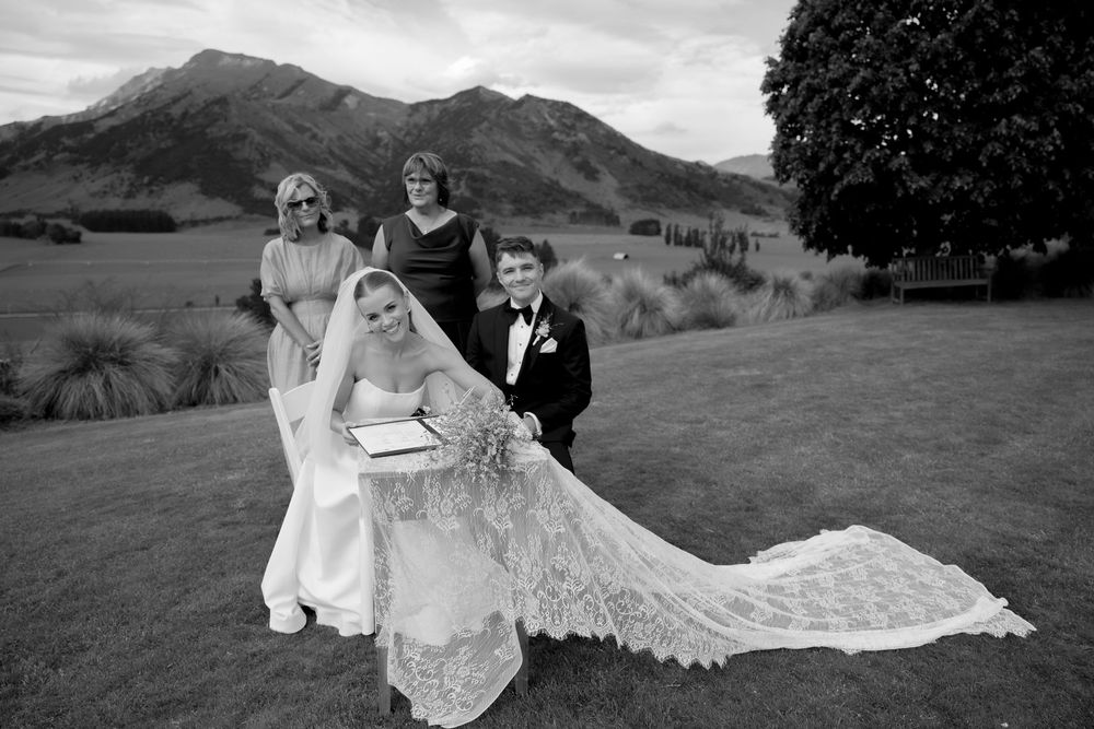 Wedding party poses with bride and groom signing register against scenic mountain backdrop.