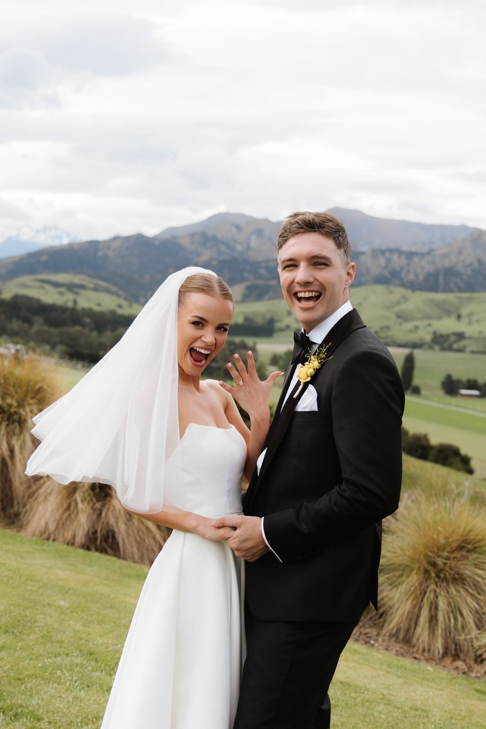 Excited bride and groom showing wedding rings while laughing with scenic mountain valley in the background.