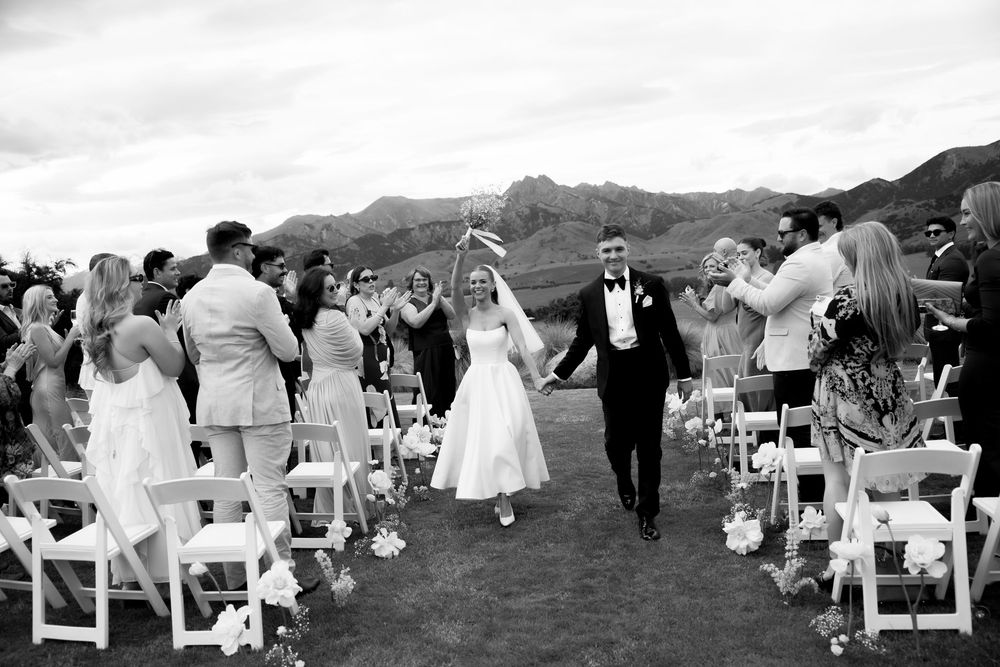 Black and white photo of newlyweds celebrating recessional with cheering guests and mountain backdrop.