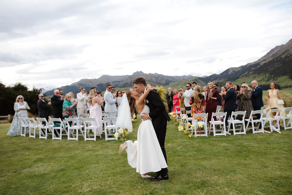 Bride and groom share first kiss at outdoor mountain wedding ceremony with guests applauding.