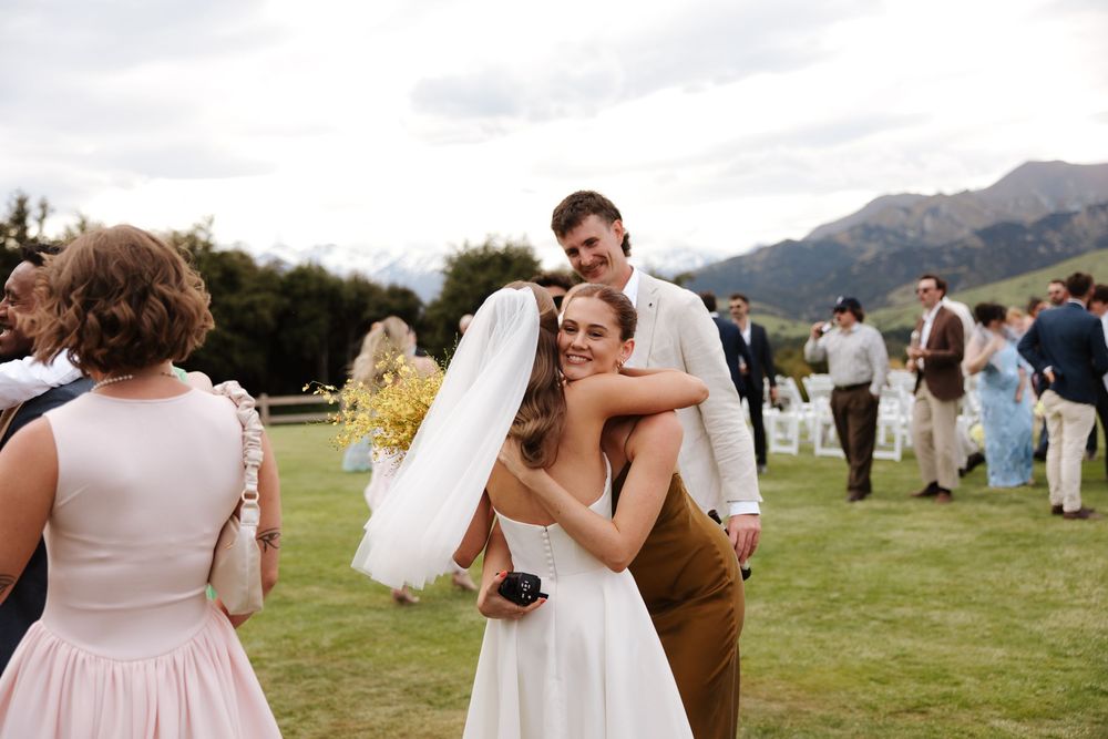 Bride hugging a wedding guest on a green lawn with mountains and wedding party visible in background.