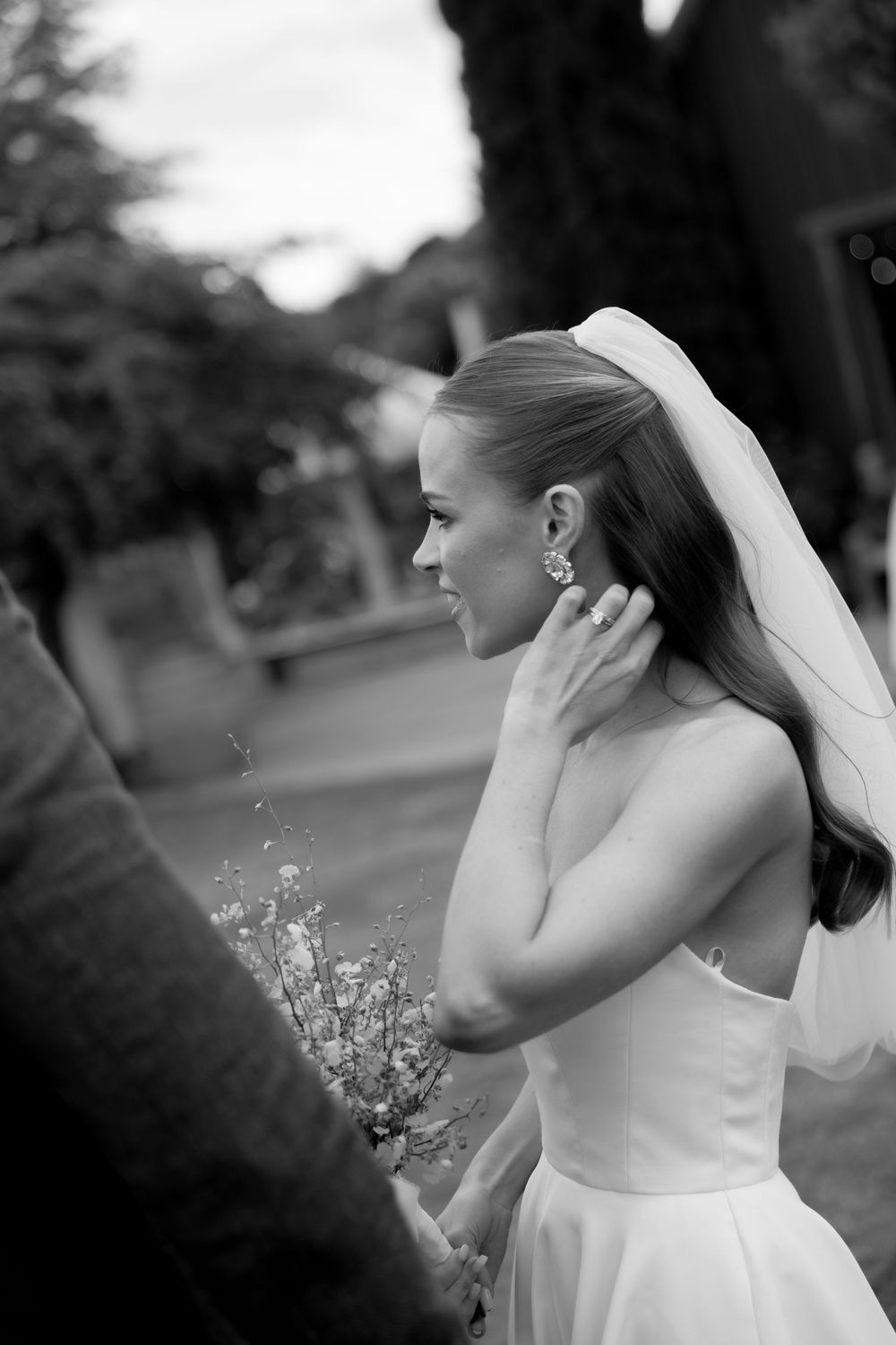 Black and white photo of a smiling bride in a strapless gown and veil, touching her earring outdoors.