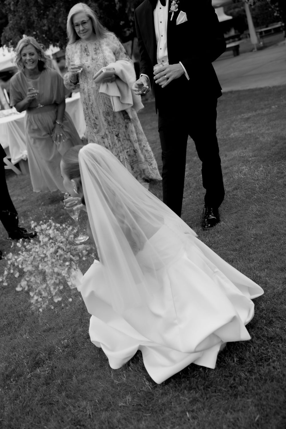 Black and white aerial view of bride in flowing veil and gown with wedding guests on grass at outdoor reception.