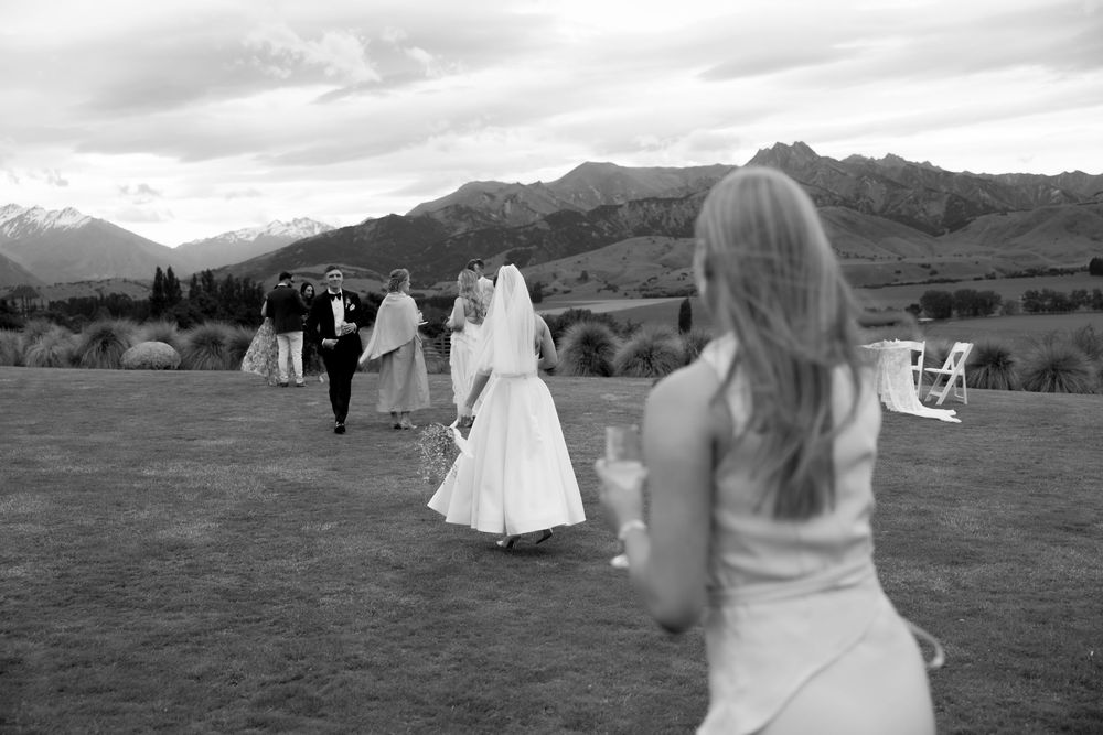 Black and white photo of wedding party walking across green lawn toward mountain backdrop.