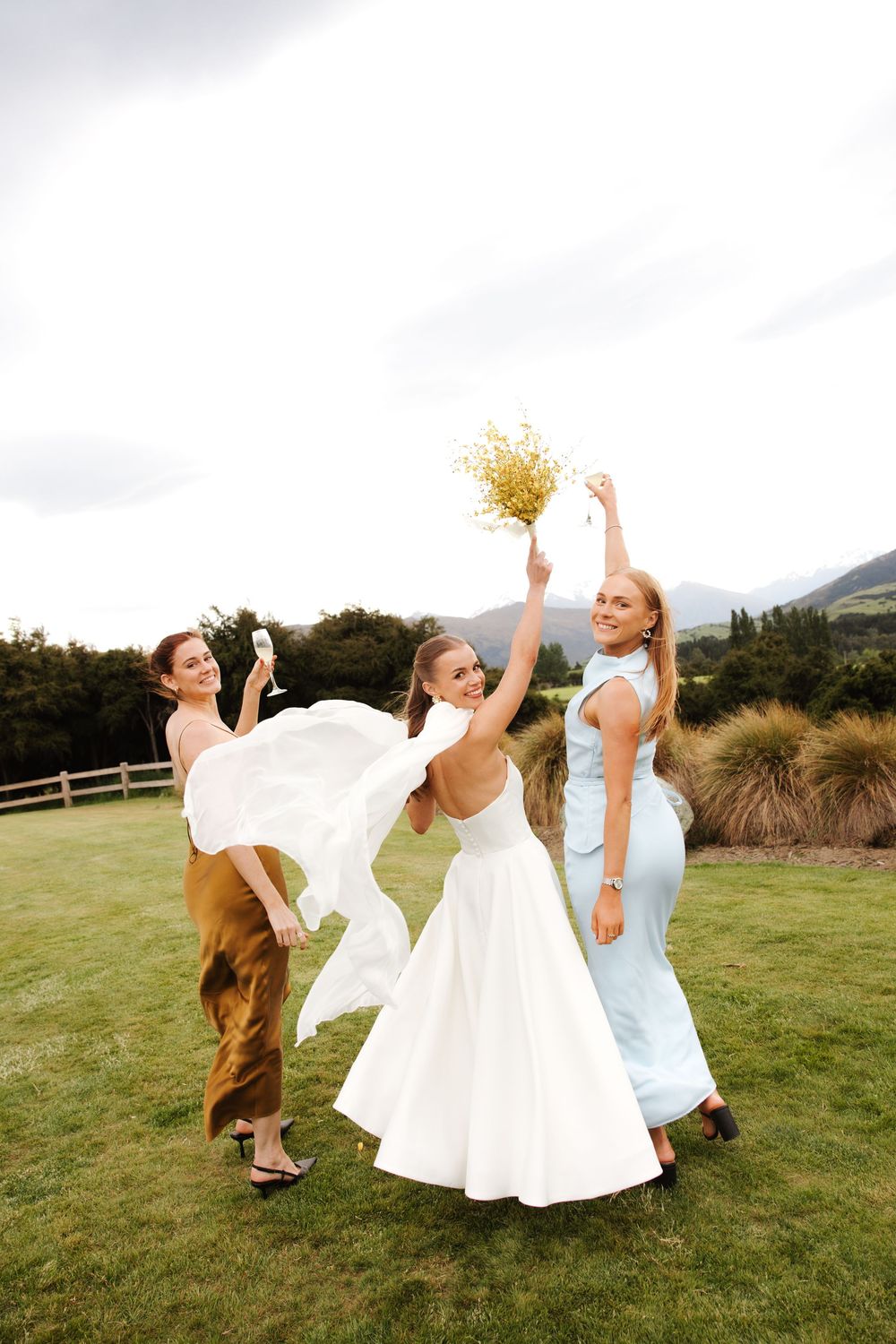 Bride in white backless gown celebrates with two bridesmaids on green lawn, mountains in background.
