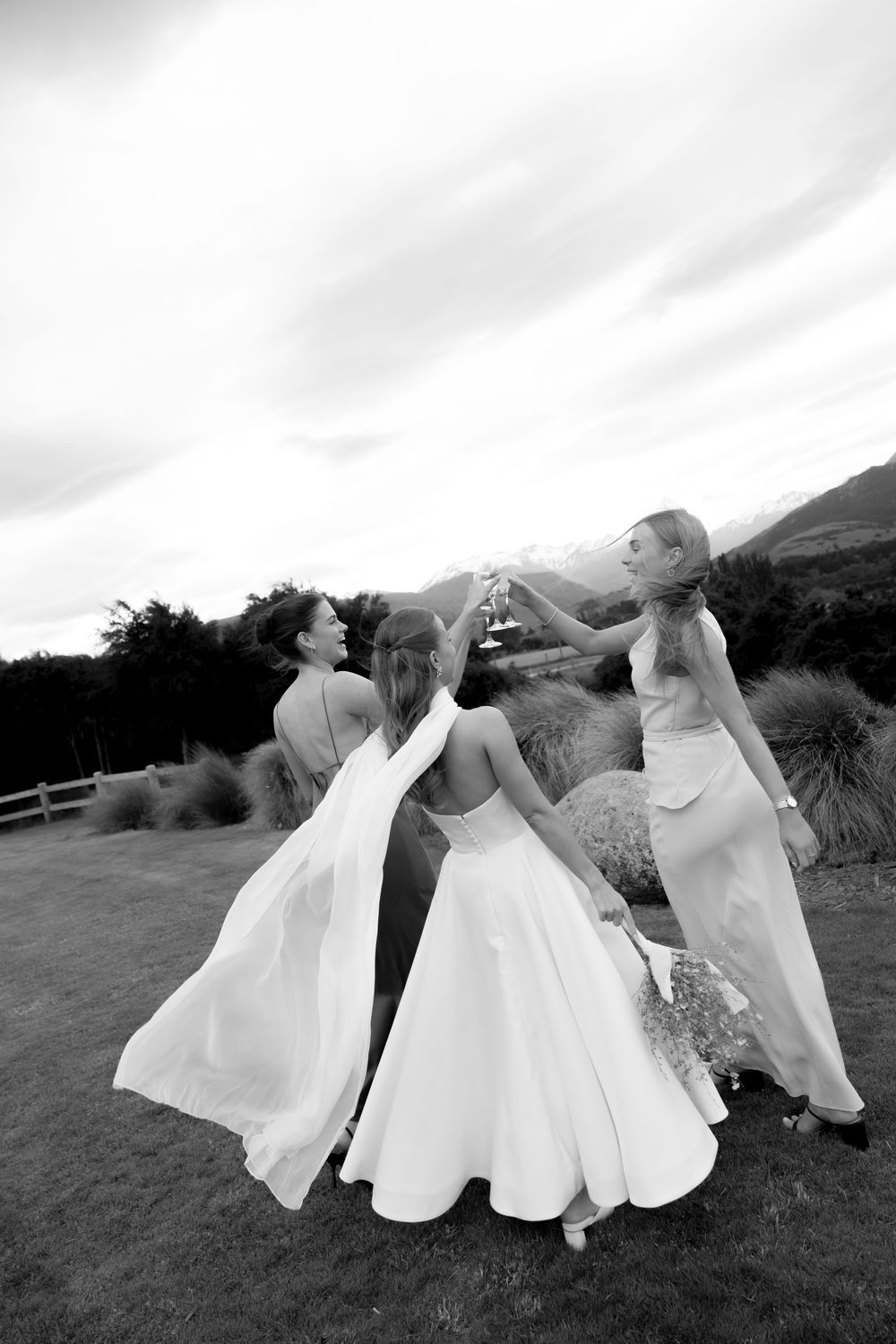 Black and white image of bride and two bridesmaids toasting champagne glasses outdoors at wedding.