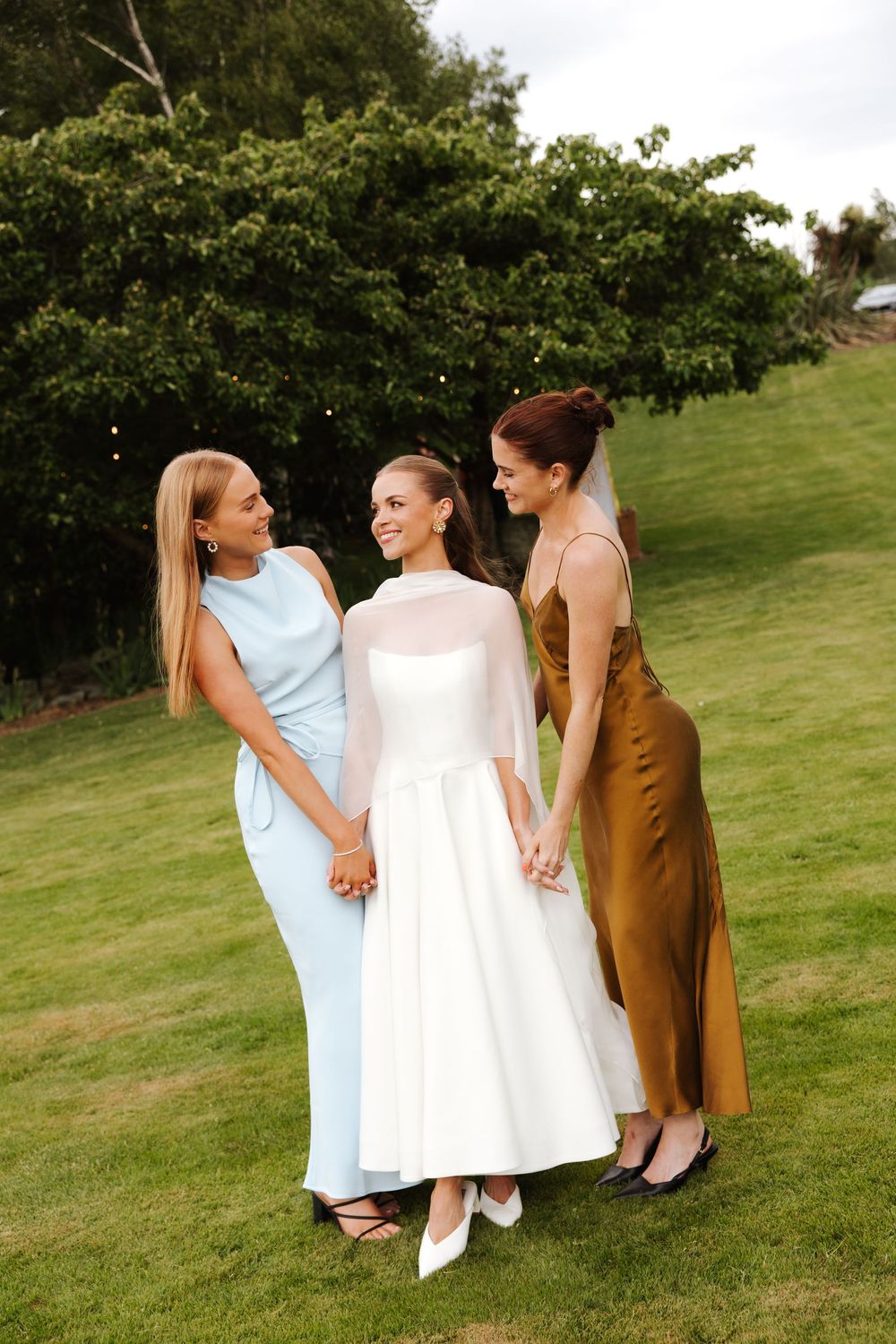 Bride in white dress holding hands with two bridesmaids in blue and gold gowns on a garden lawn.