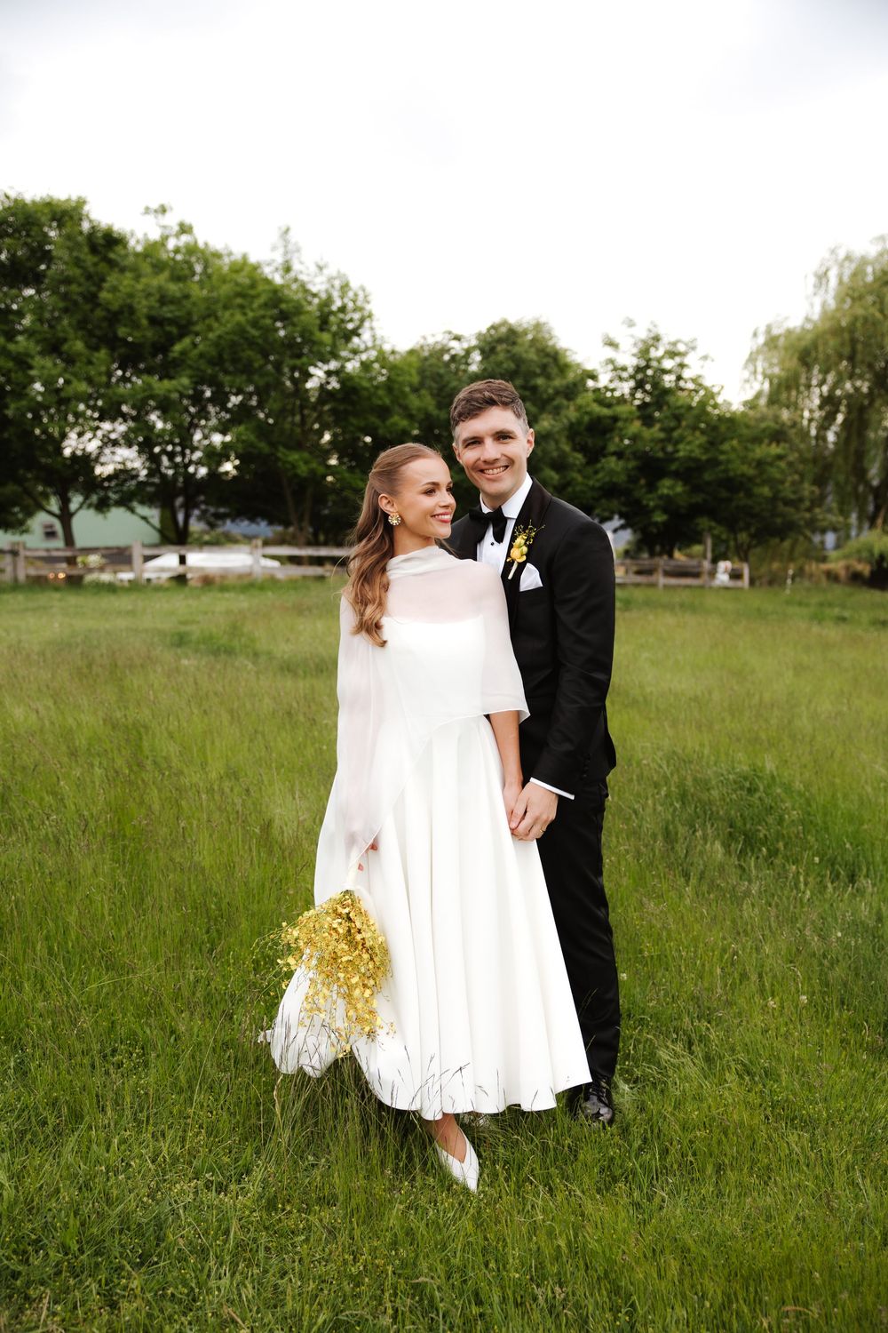 Smiling bride and groom pose together in green field, bride in white midi dress holding yellow bouquet.