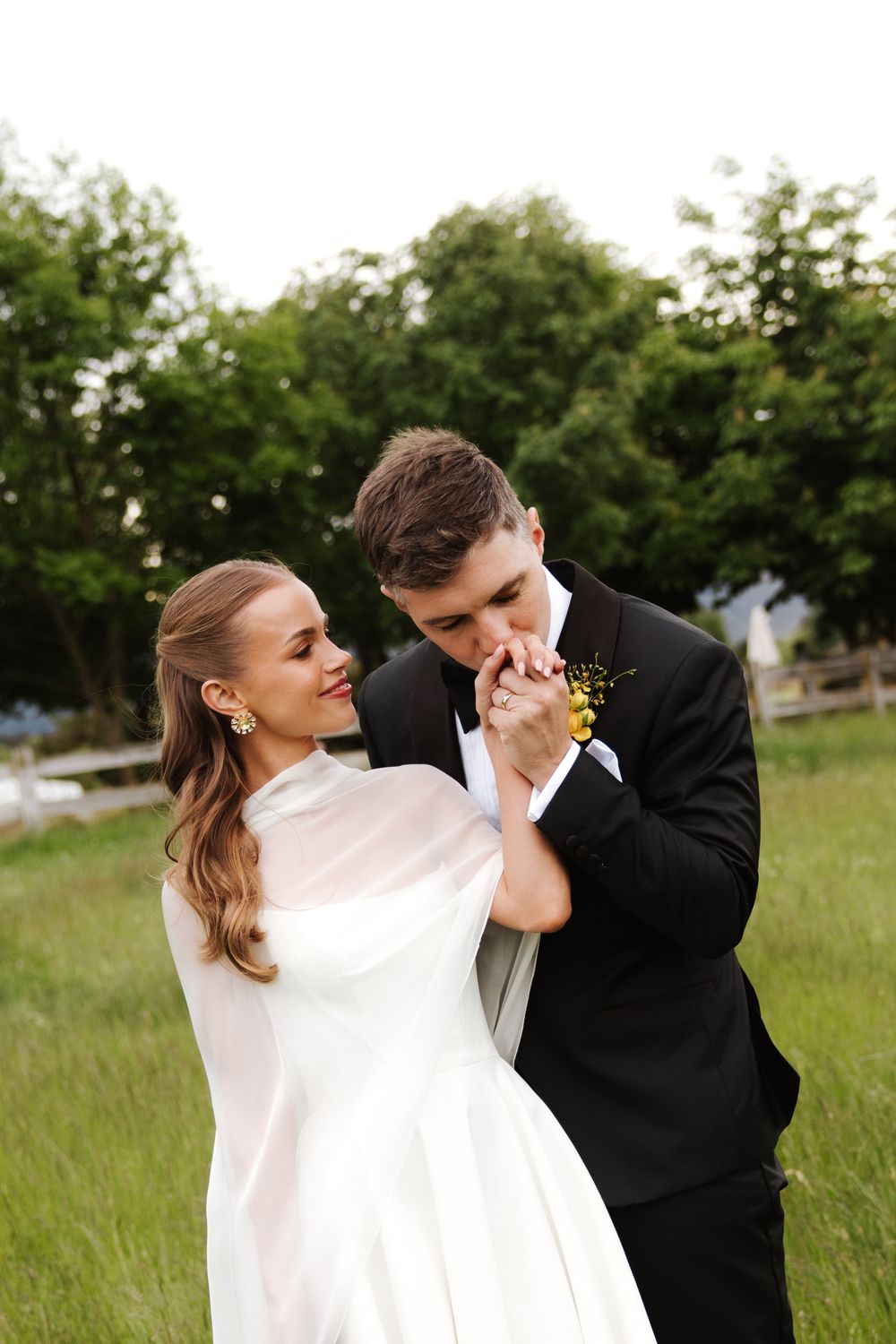 Groom in black tuxedo kissing brides hand while she smiles, standing in green field at outdoor wedding.
