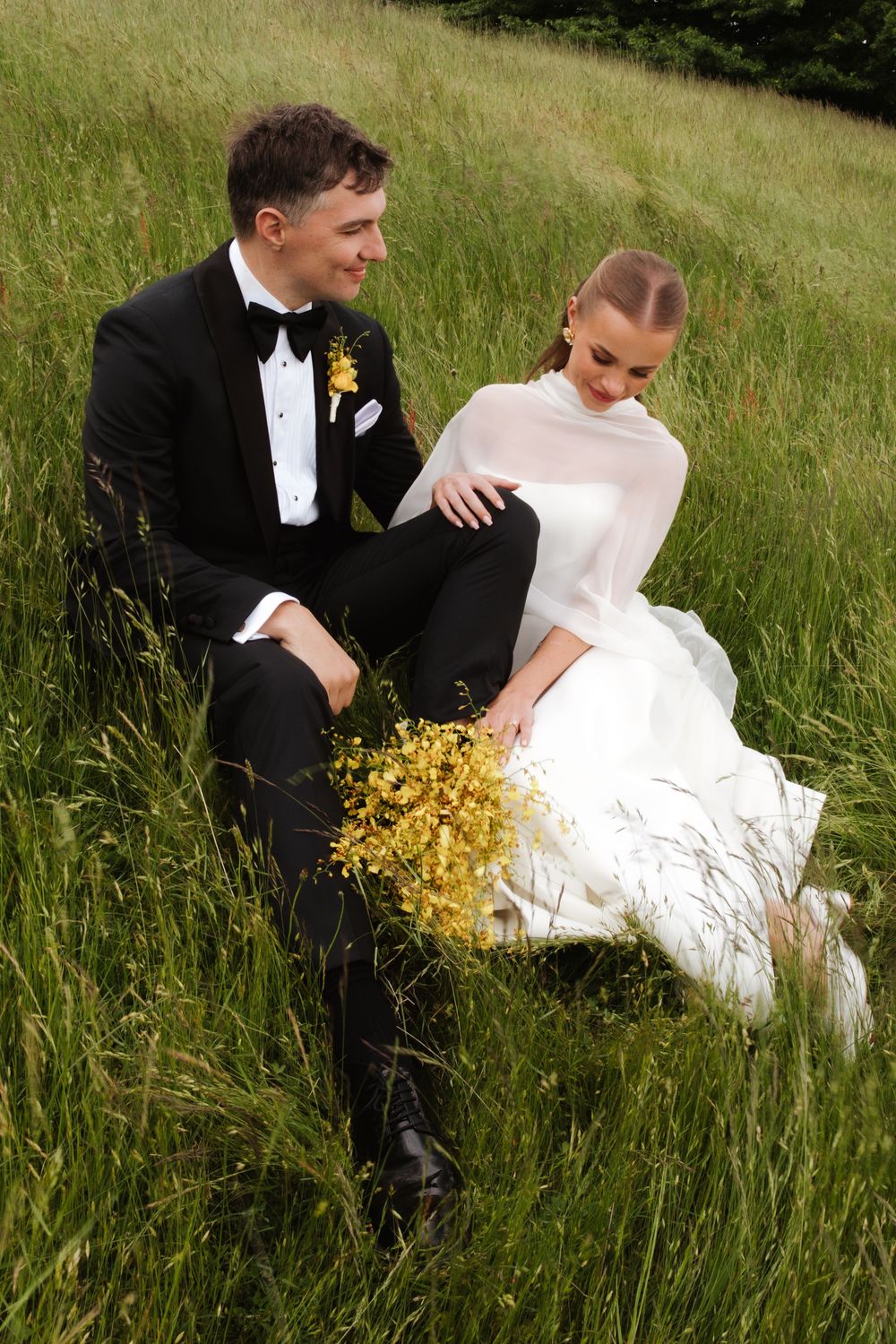 Bride and groom sitting together in tall green grass holding a yellow wildflower bouquet on their wedding day.