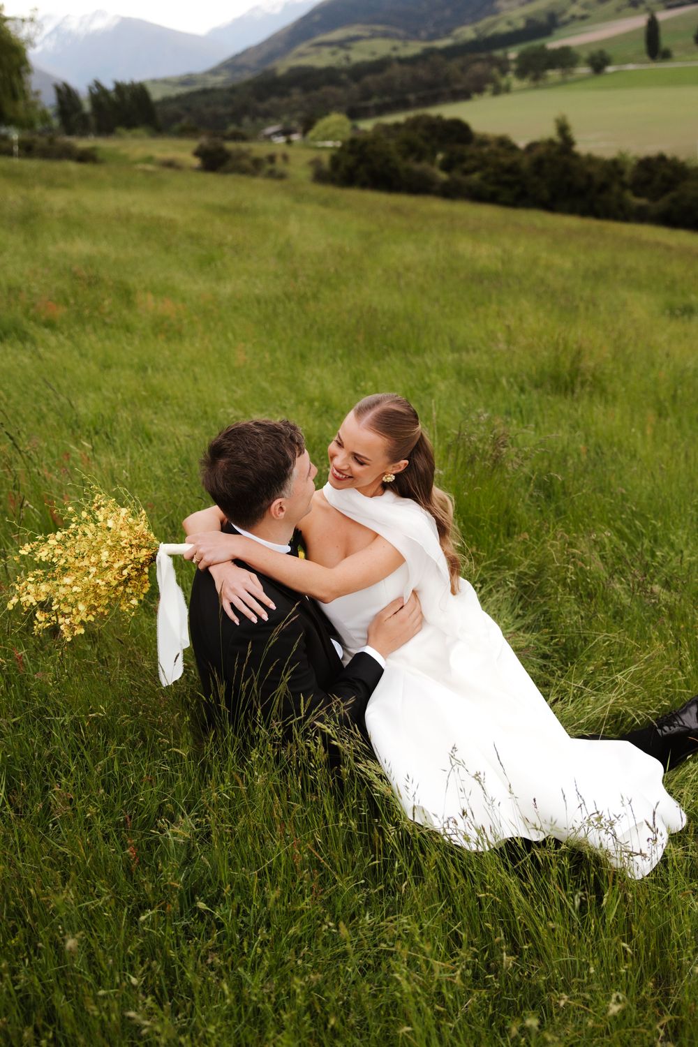 Bride and groom sit together in lush green field, sharing a joyful moment with yellow bouquet nearby.
