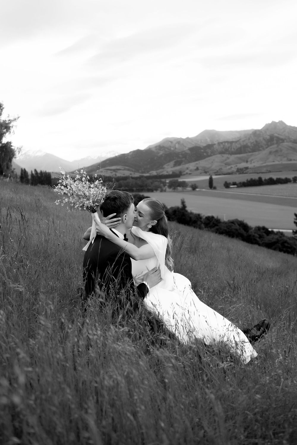 Black and white photo of bride and groom kissing while sitting in tall grass with mountain landscape behind them.