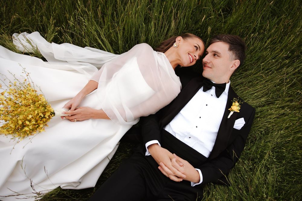 Bride and groom lying on green grass holding hands, with a yellow wildflower bouquet beside them.