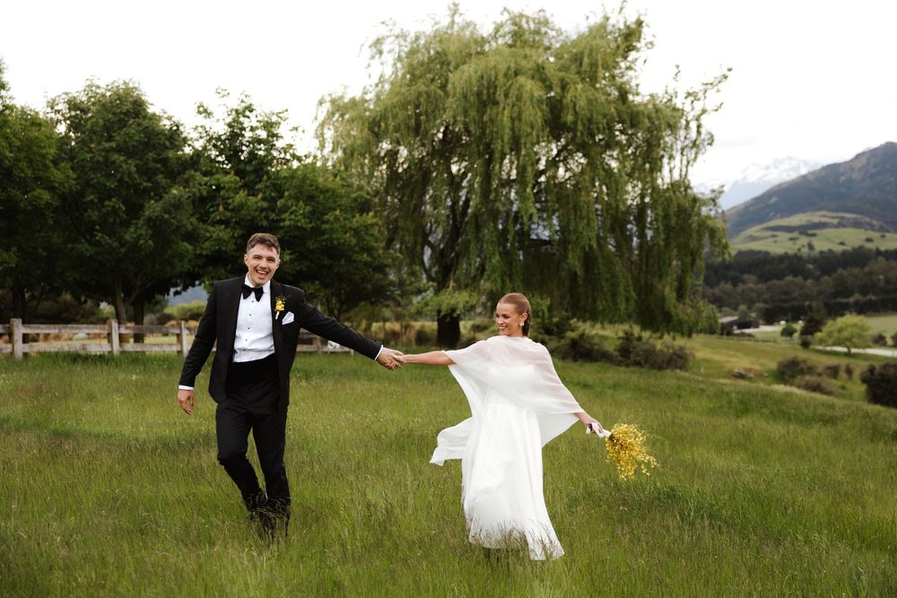 Bride and groom walking hand in hand through a green meadow with a willow tree and mountains behind them.
