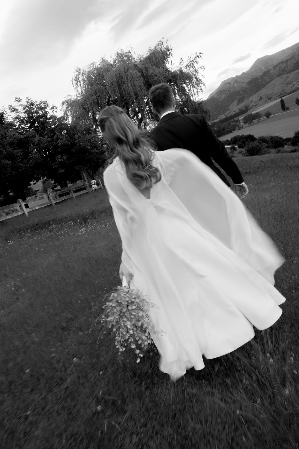 Black and white rear view of bride in flowing veil and groom walking hand in hand across green field.