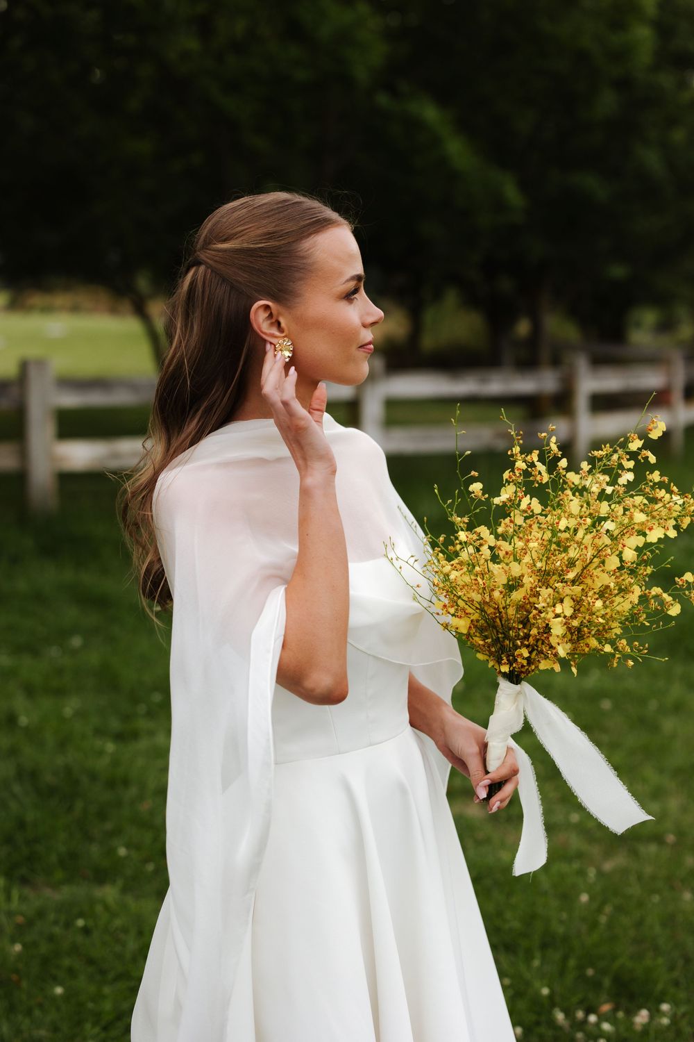 Bride in white cape gown holds yellow wildflower bouquet outdoors near rustic wooden fence.