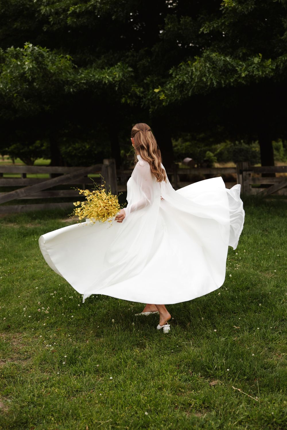 Bride in flowing white dress twirling with yellow bouquet in green garden setting.