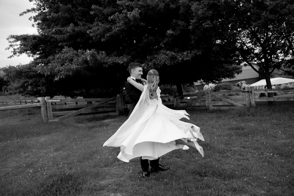 Black and white photo of groom twirling bride in flowing white dress on green lawn.