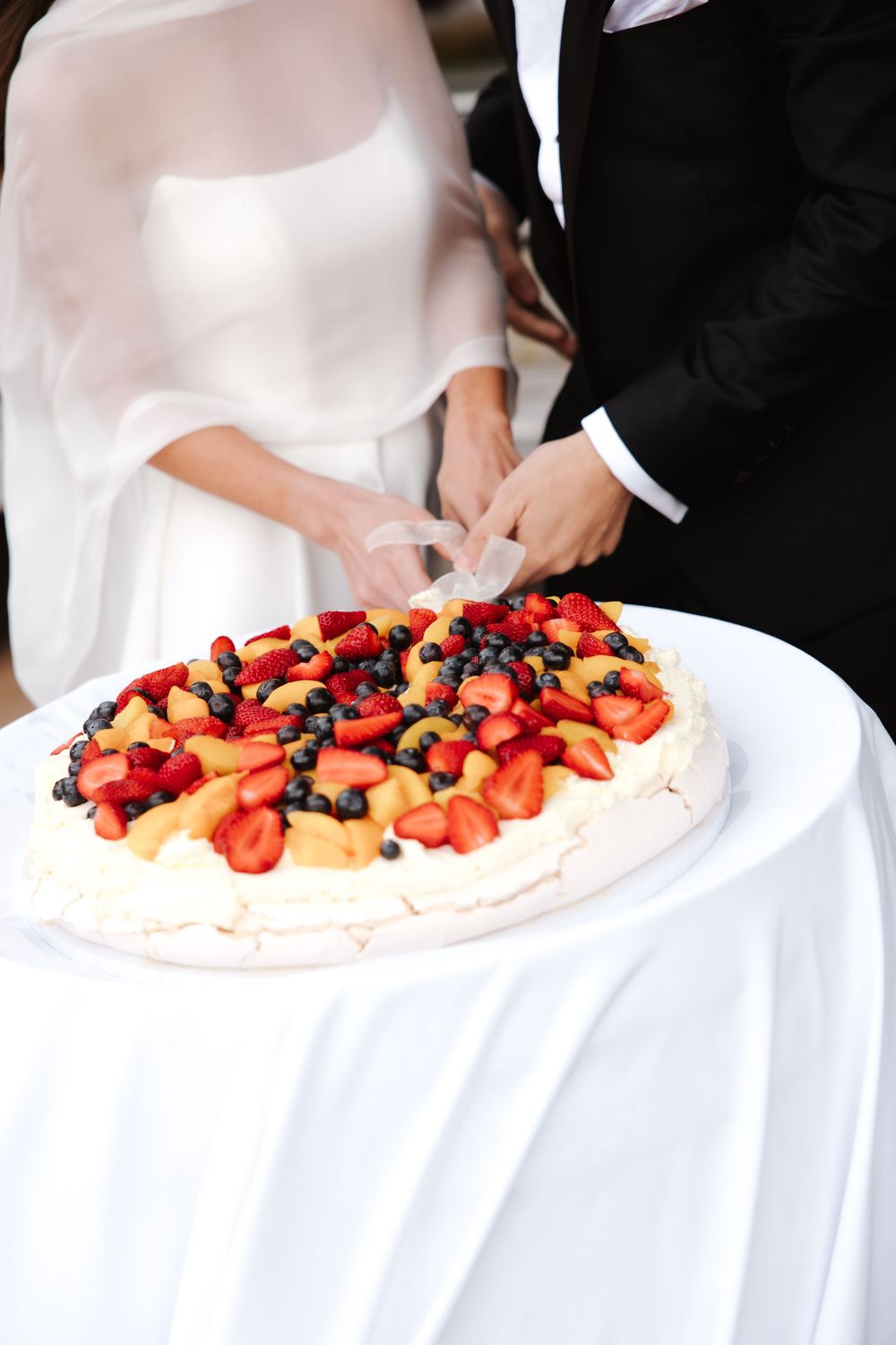 Close-up of bride and groom cutting pavlova wedding cake topped with colorful fresh fruits.