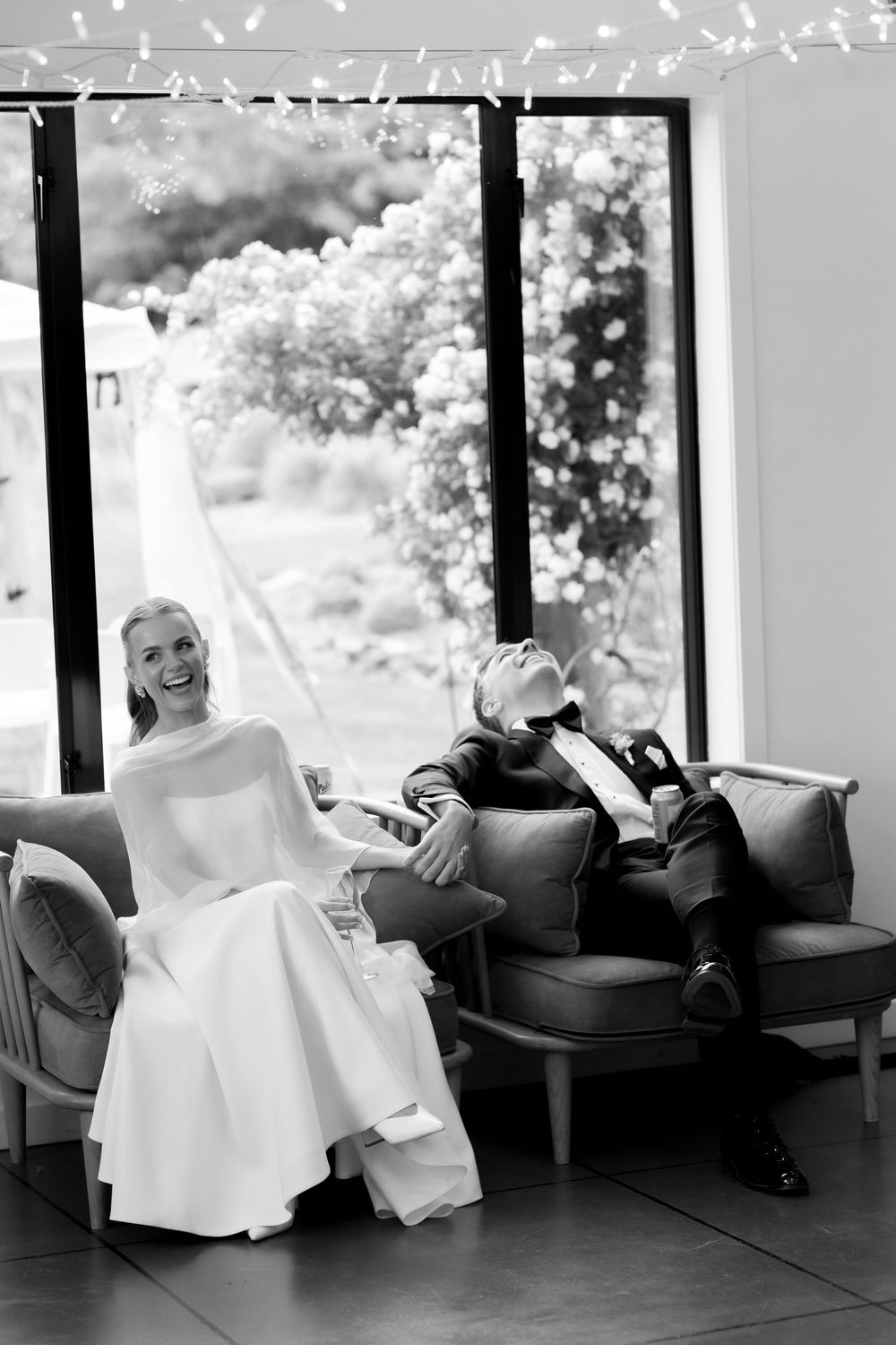 Black and white photo of bride and groom laughing on a sofa with fairy lights and garden views.