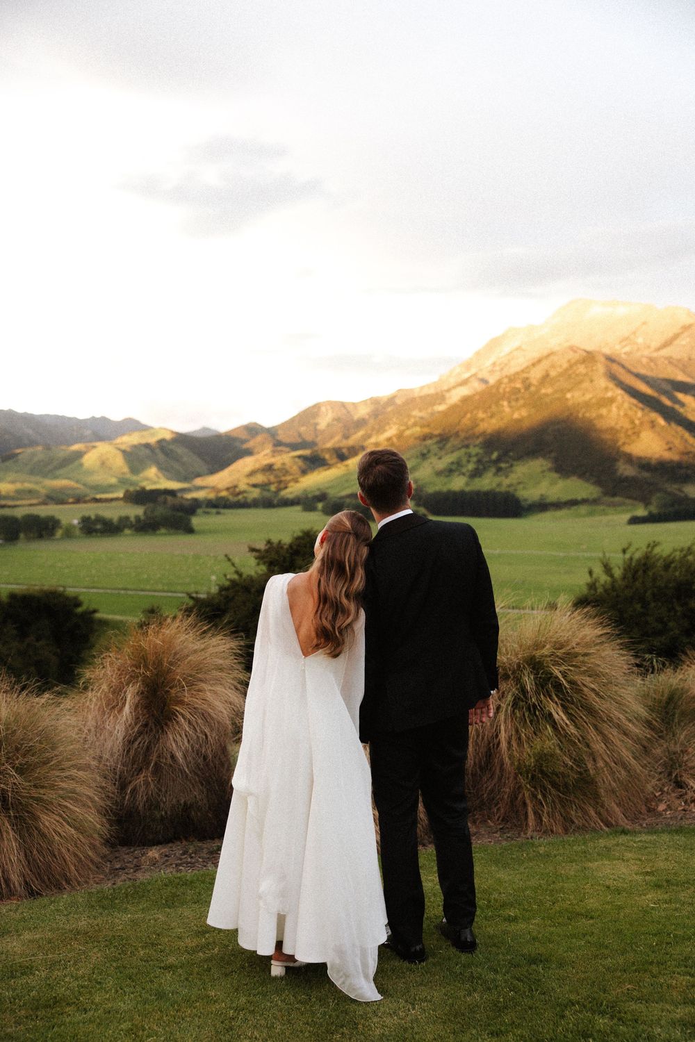 Bride and groom stand together gazing at stunning mountain valley landscape at golden sunset.