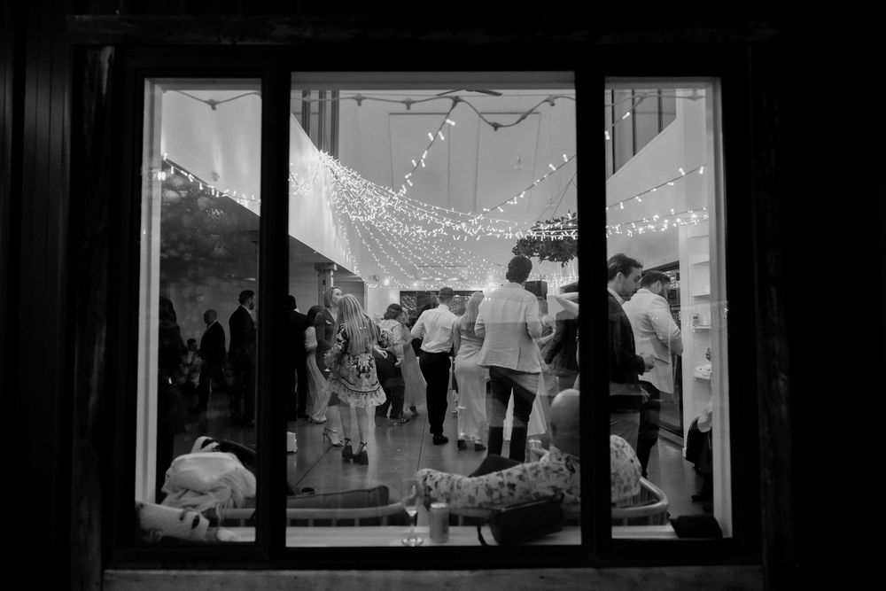 Black and white view through window of wedding reception guests dancing under fairy lights.