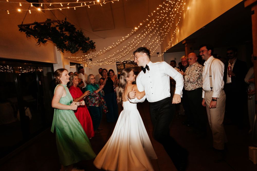 Bride and groom share first dance surrounded by guests under warm string lights at elegant wedding reception.