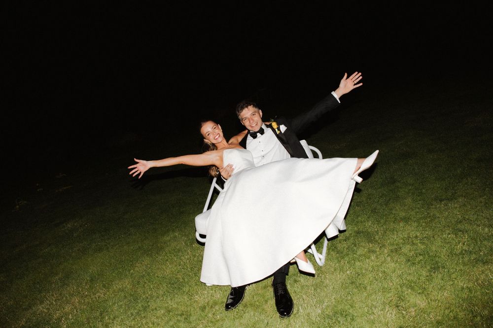 Groom lifts smiling bride in white gown at night, both posing with arms outstretched on grass.