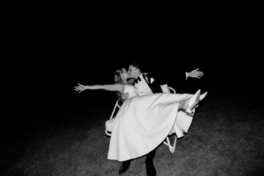 Black and white photo of groom lifting bride in wedding attire outdoors at night.