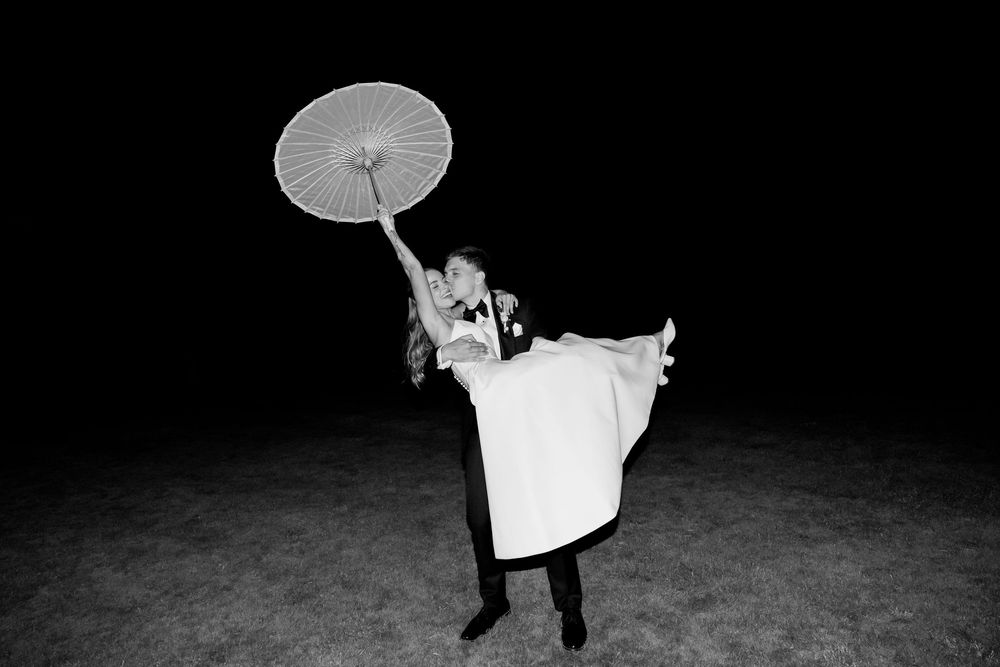 Black and white night photo of groom lifting bride while she holds a parasol umbrella at wedding reception.