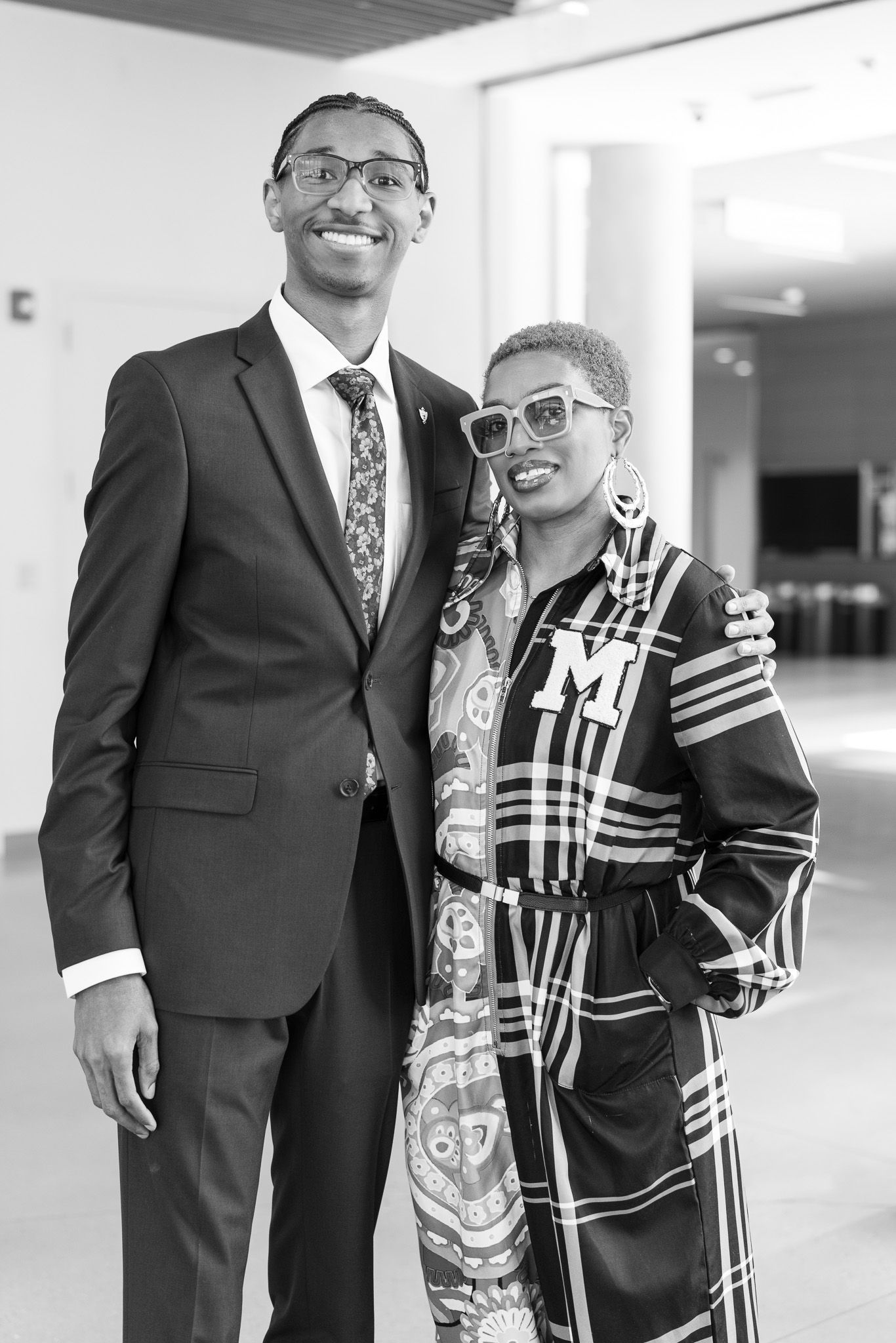 Black and white photo of two people posing together, man in suit and tie, woman in plaid outfit and sunglasses.