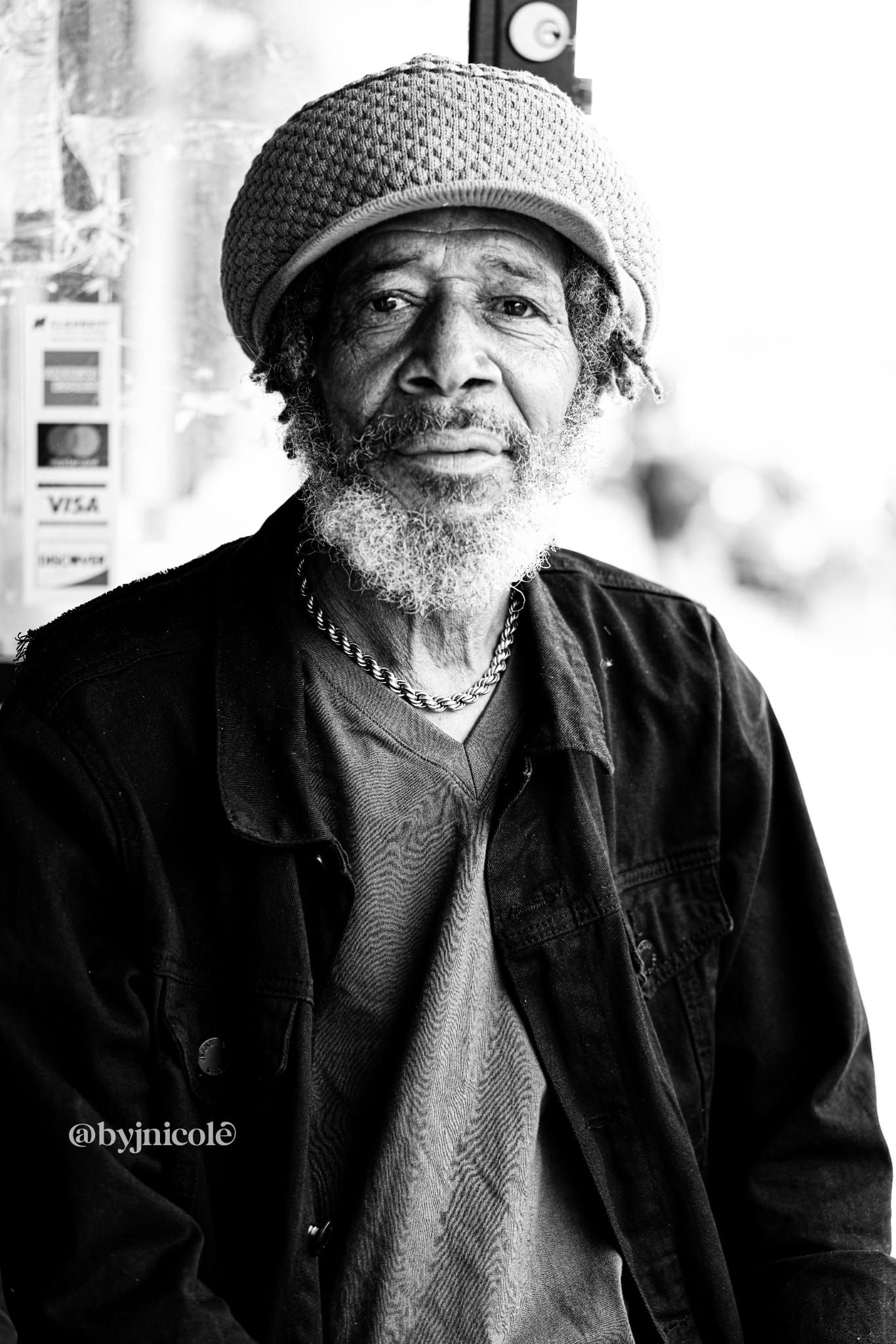 Black and white portrait of an elderly man wearing a knit cap and jacket, with a beard and chain necklace.