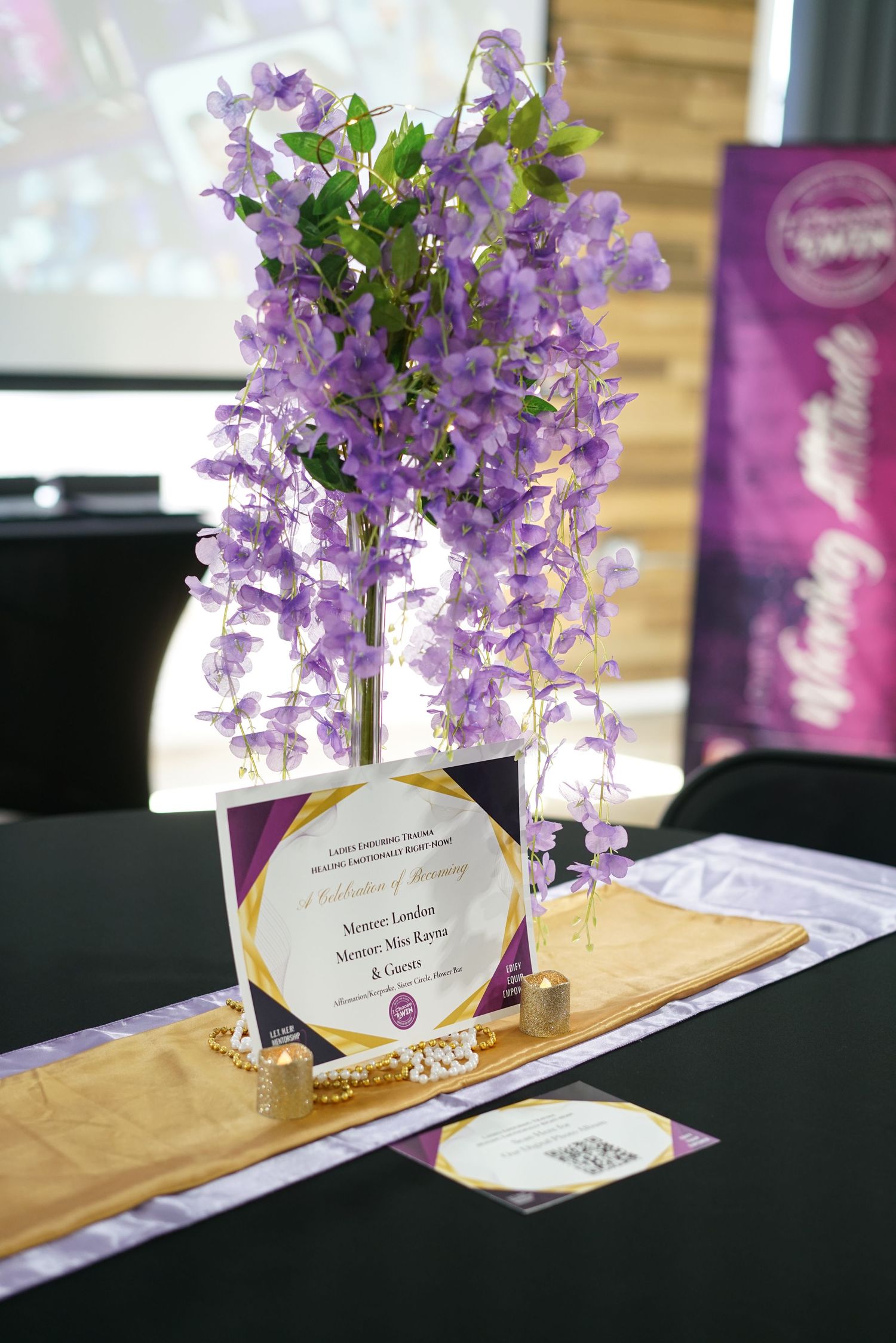 Elegant event table centerpiece with purple wisteria flowers, gold runner, and celebration signage.