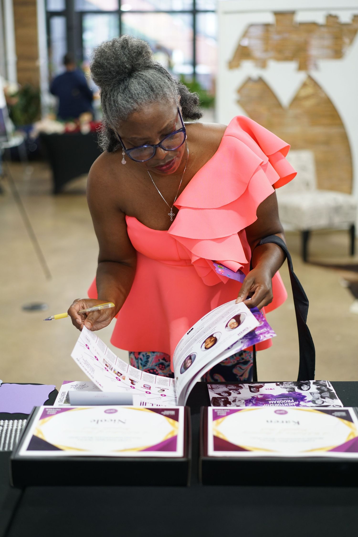 Woman in coral ruffled top reviewing a magazine or catalog at a vendor table with purple gift boxes.
