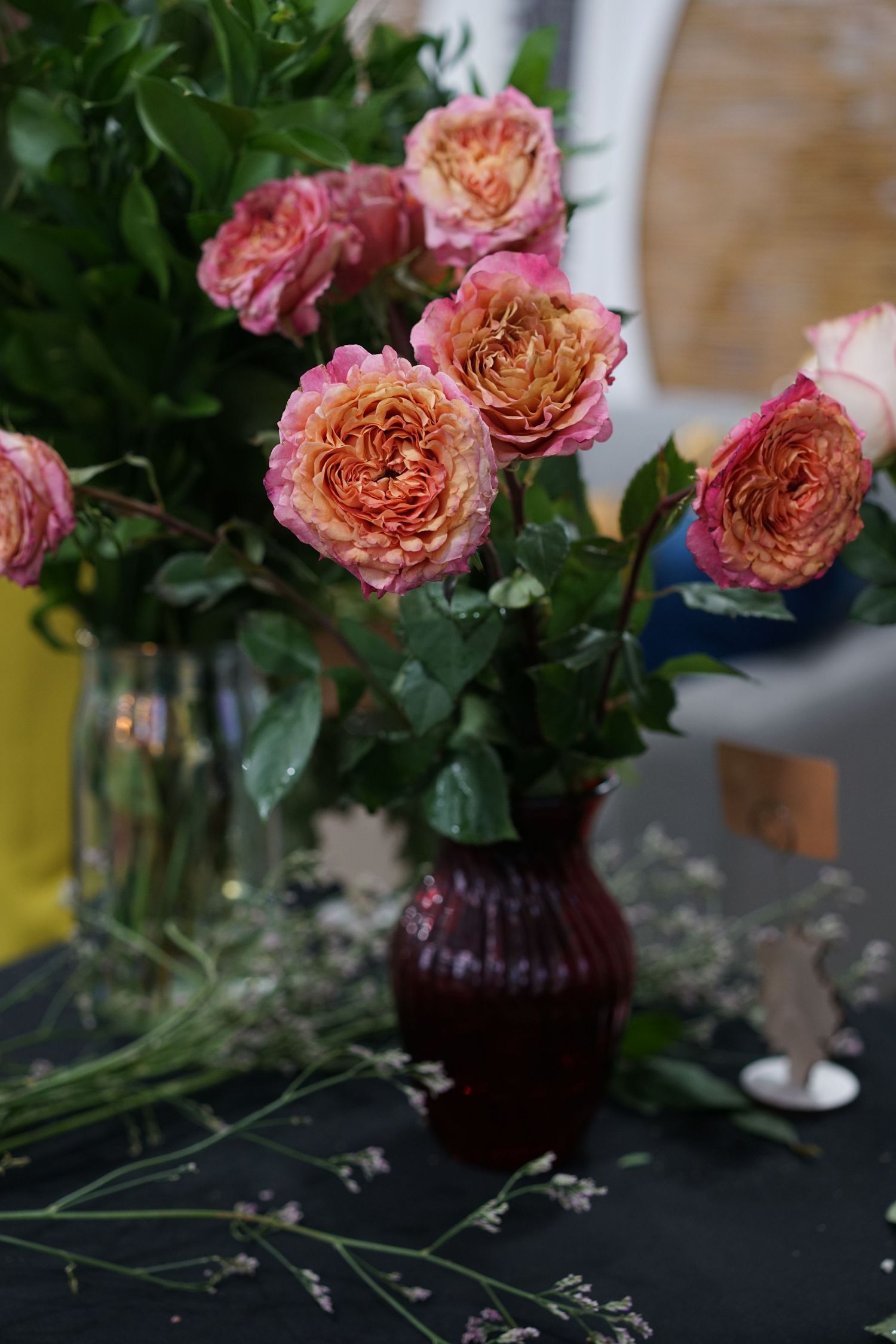 Dark red vase with pink and orange garden roses surrounded by greenery on a dark table.