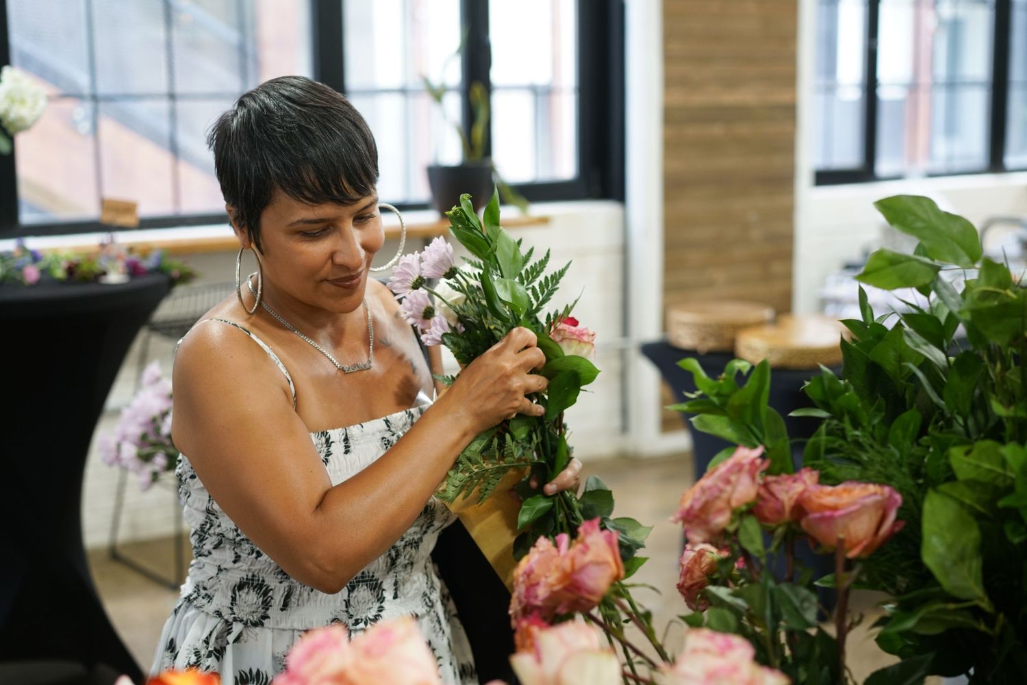 Woman in floral dress carefully arranging a bouquet of pink flowers at an indoor floral workshop.