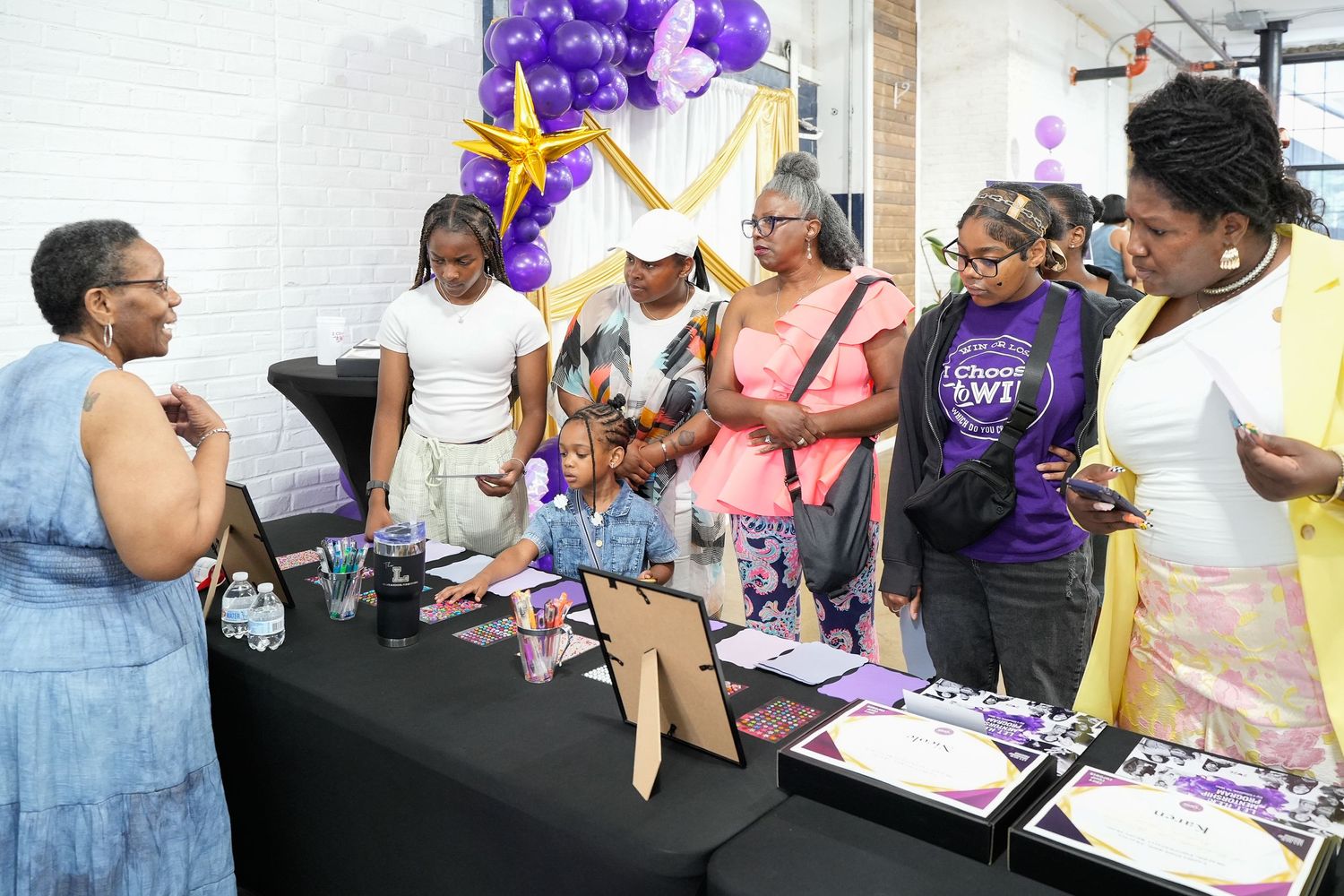 Group of Black women and girls gathered around a table with purple decorations and balloons at a community event.