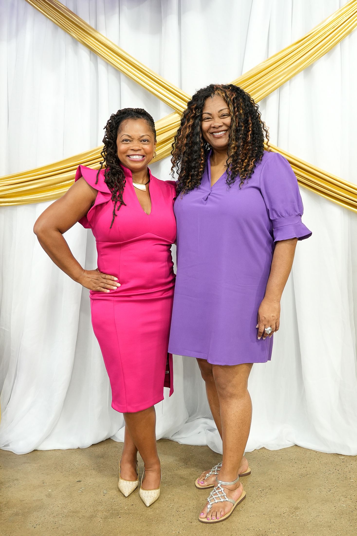 Two women smiling in pink and purple dresses posing in front of a white and gold backdrop.