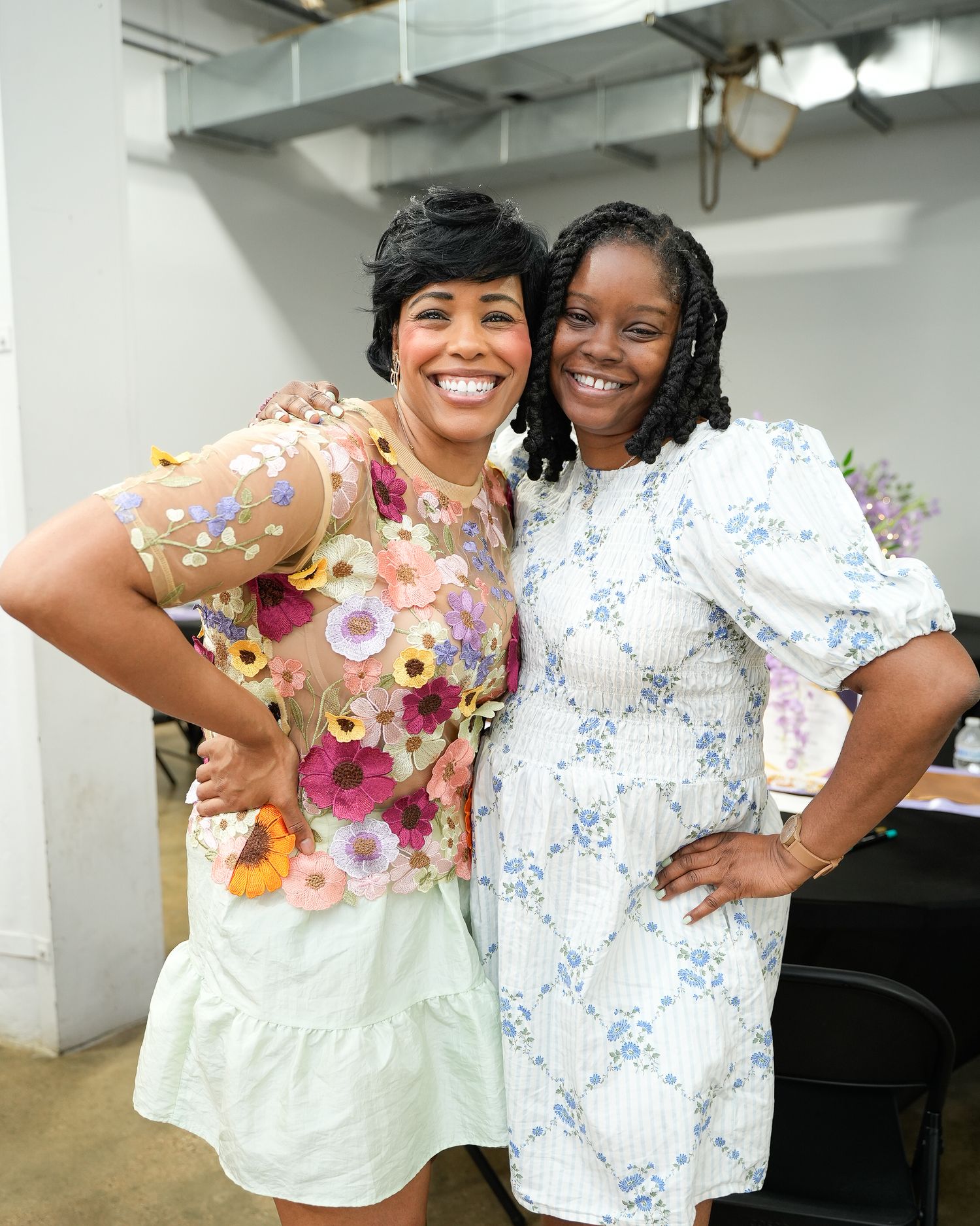 Two women smile and pose together at an indoor event, wearing floral and blue dresses.