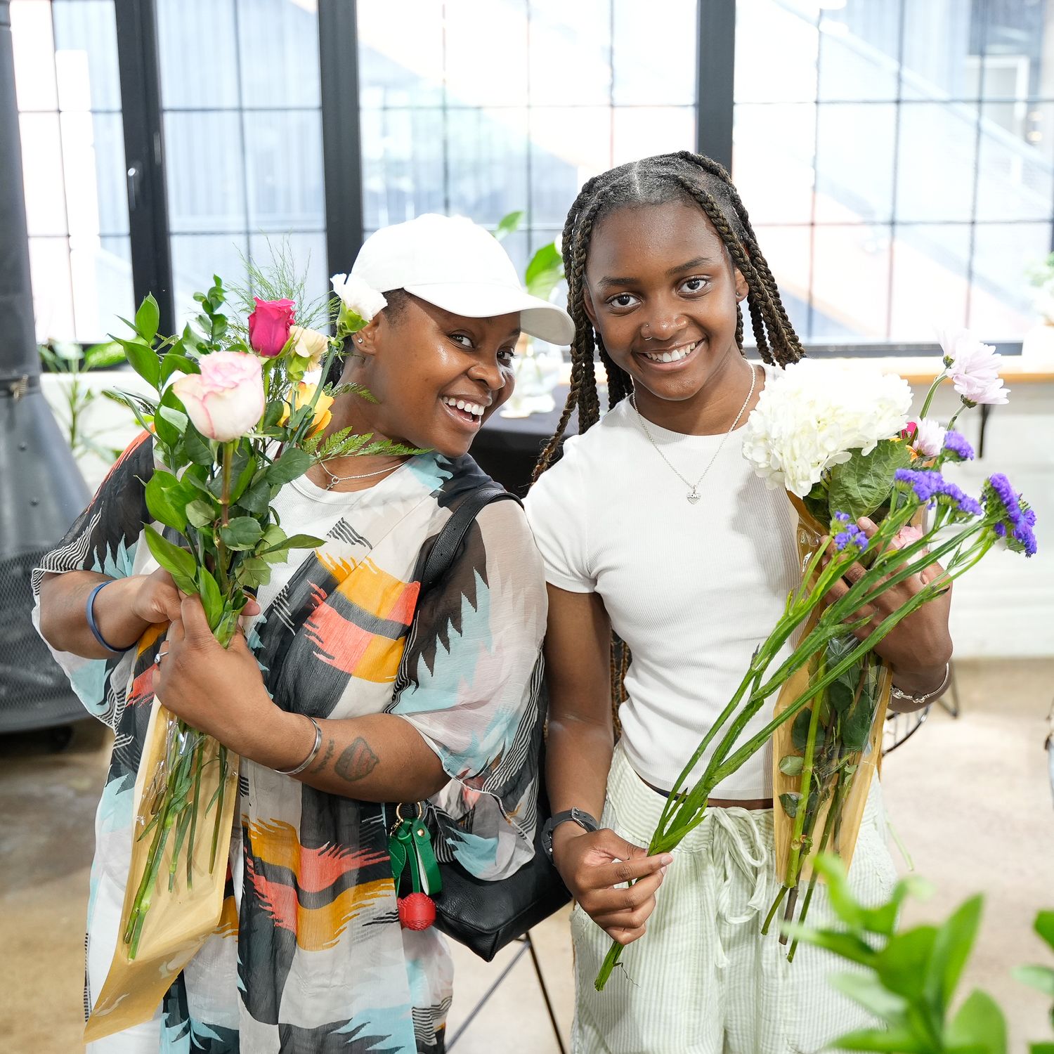 Two women smile holding colorful flower bouquets in a bright indoor space, one wearing a white cap and patterned top.
