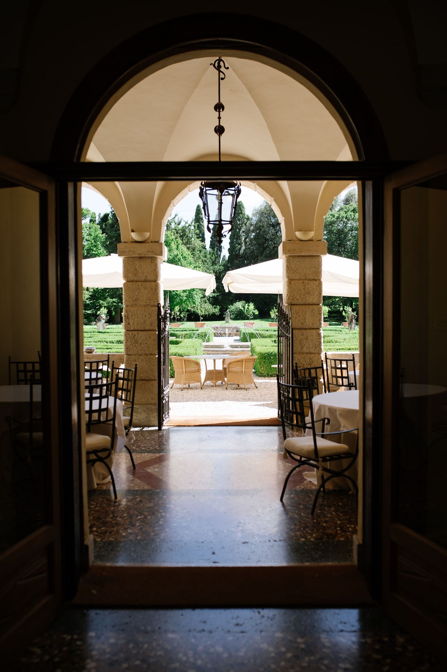 Elegant arched doorway opens to a sunlit Italian villa courtyard with wrought iron chairs and manicured garden beyond.