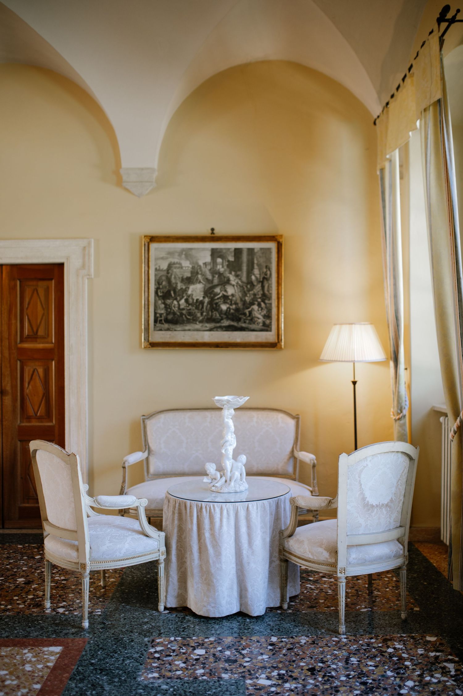 Elegant Italian sitting room with Louis XVI chairs, draped table, white sculpture, arched alcove, framed artwork, and warm floor lamp.