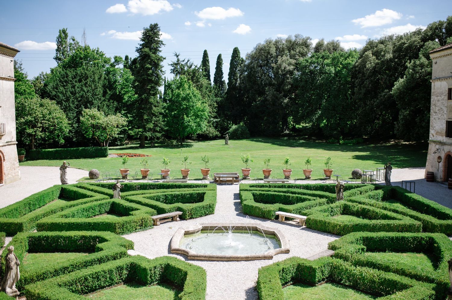 Formal Italian garden with geometric boxwood hedges, central fountain, and lush green trees under a bright blue sky.