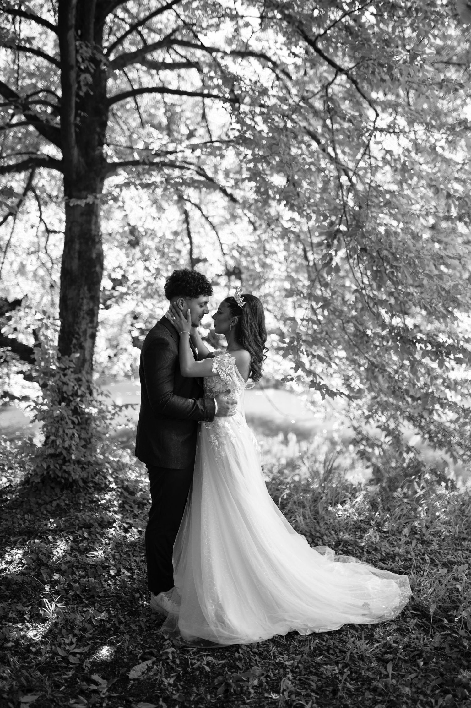 Black and white photo of a bride and groom embracing under trees in a romantic outdoor wedding portrait.
