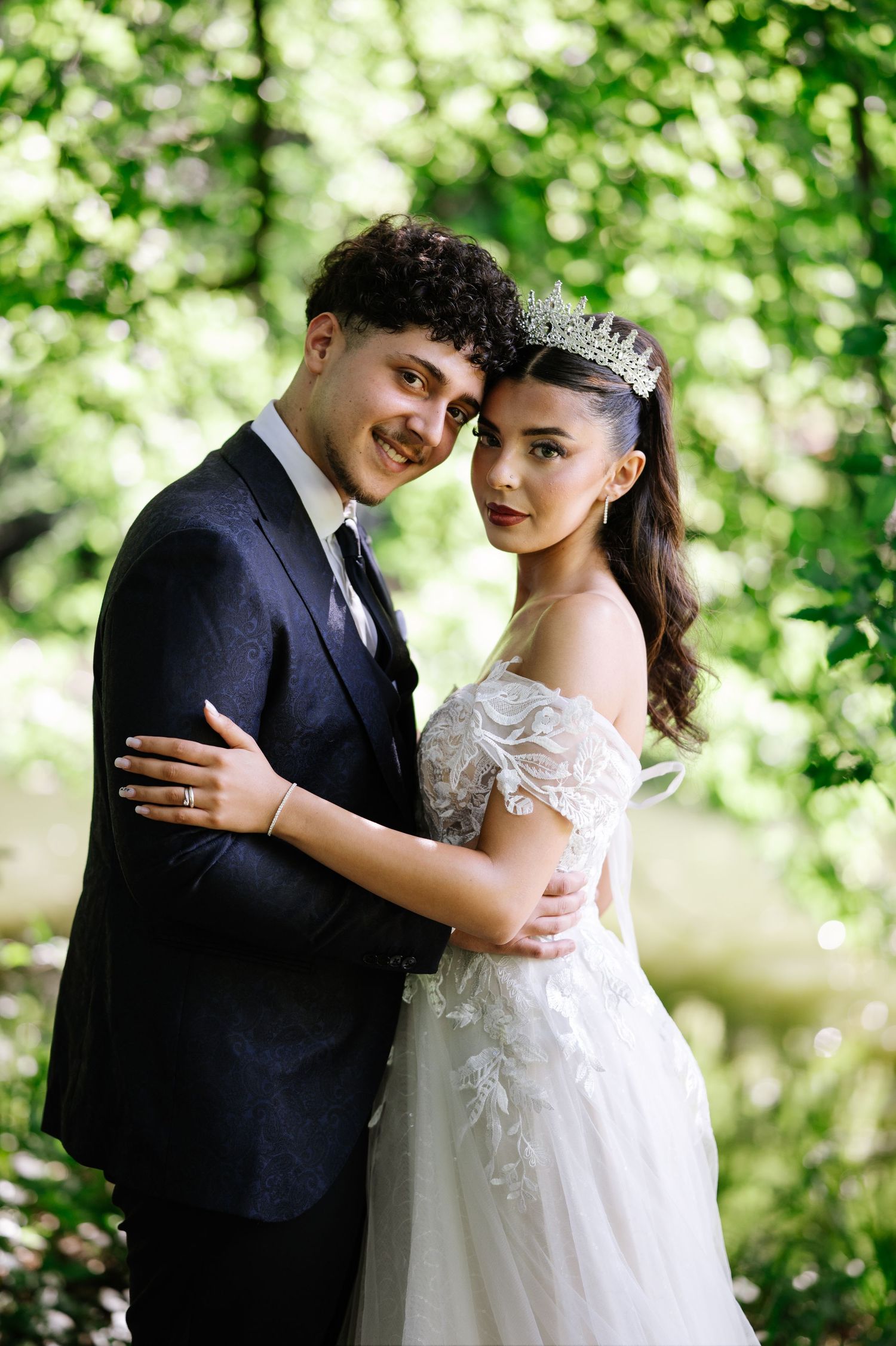 Bride in lace off-shoulder gown and tiara embraces groom in dark suit outdoors among lush green trees.