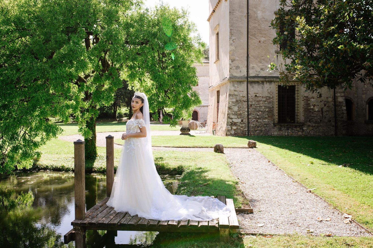 Bride in white gown and veil stands on wooden dock by pond near historic stone building surrounded by lush green trees.